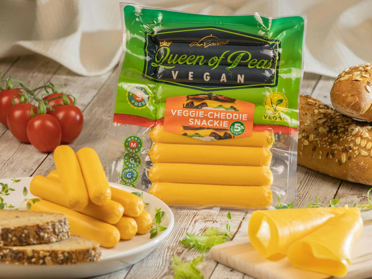 Veggie-Cheddie Vegan Snackie in packaging with slices of snack on table, surrounded by bread, tomatoes, and herbs.